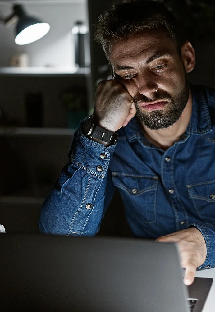 Tired young man in a denim shirt resting his head on his hand while looking at a laptop screen in a dimly lit room.